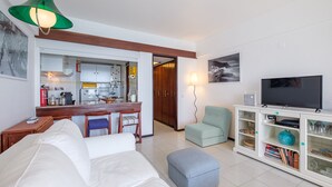 A living room with a sofa, ottoman, TV stand, and a dining area with a kitchen in the background. - Direct & Private beach access. 30 km from Lisbon (Sesimbra)