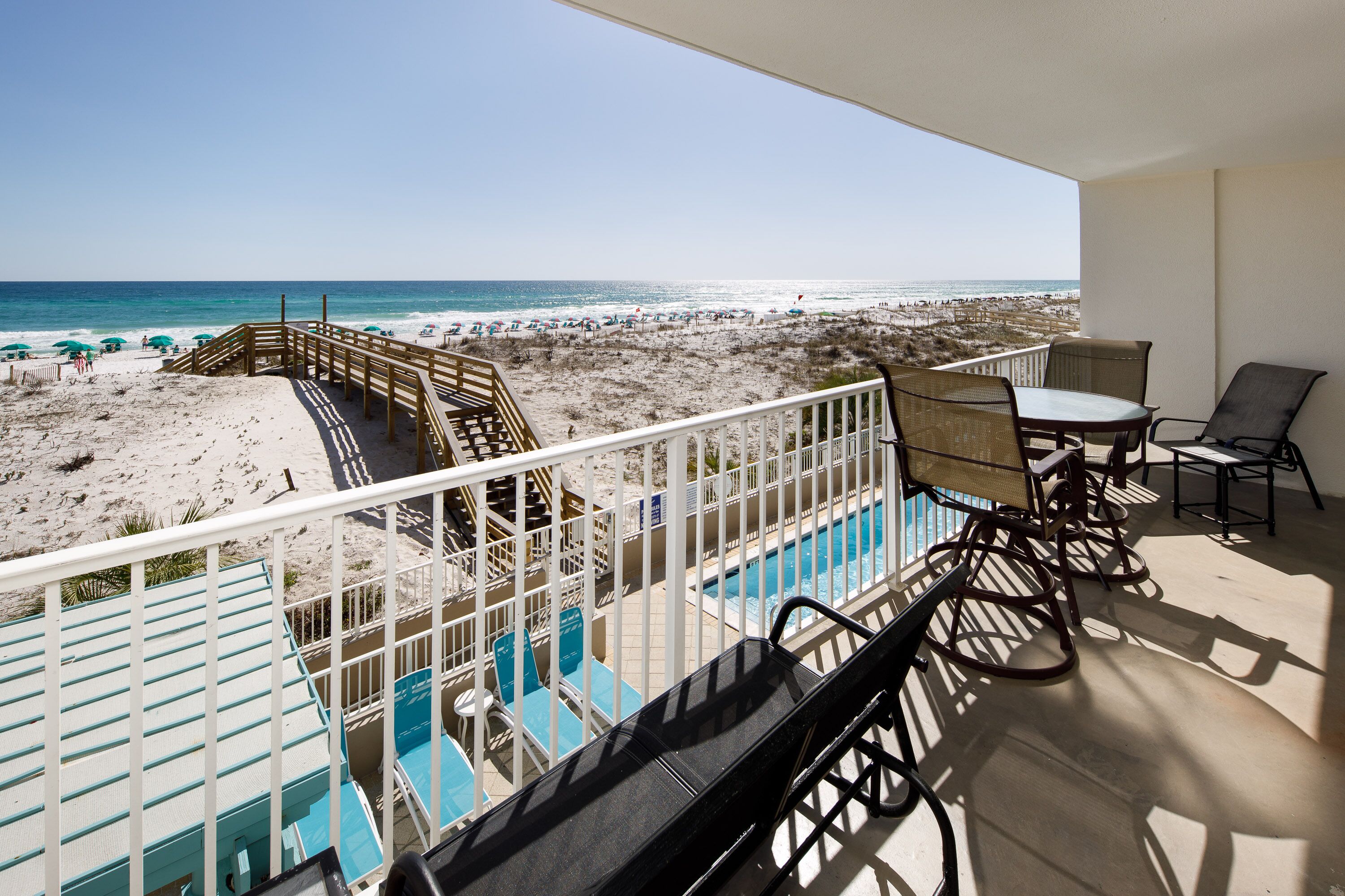 A balcony with a view of the beach, a pool, and outdoor seating.