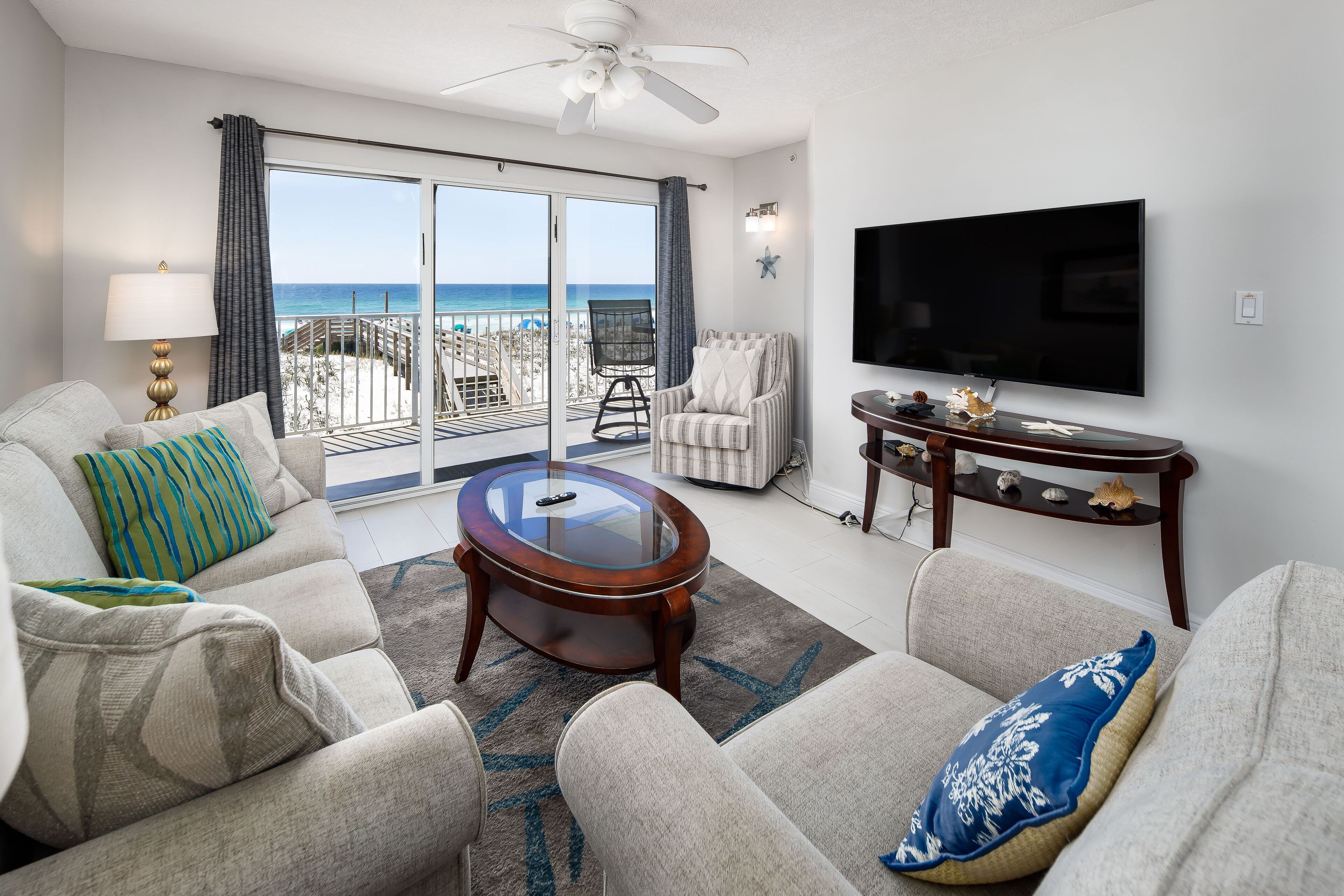 A living room with a large TV, comfortable seating, a glass coffee table, and a view of the beach.