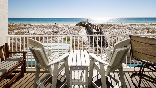 A wooden deck with white chairs and a table overlooking a beach with a wooden walkway leading to the water.