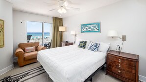 A bedroom with a bed, a dresser, a chair, a nightstand, a lamp, a painting, and a view of the beach through the window.
