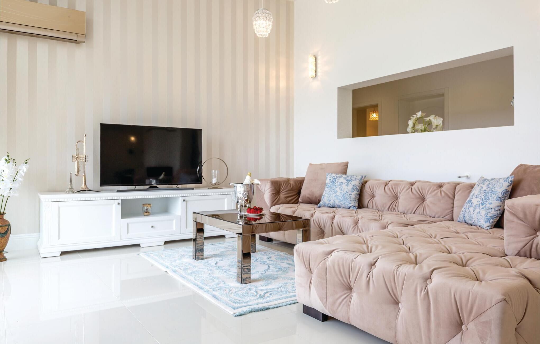 Una sala de estar moderna con un televisor de pantalla plana grande, un mueble blanco, un sofá color beige con almohadas azules y un espejo decorativo en la pared.