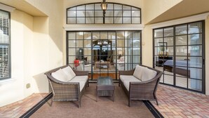 A covered patio with wicker furniture, a glass table, and a view of the interior through large windows. - Private Pool | Single Floor Home | Near Attractions (Kissimmee)