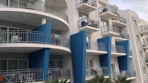 A multi-story residential building with balconies, a parking area, and a few parked vehicles. - Azure Cottage: Beautiful, modern 3 bedroom apartment with communal swimming pool (Marsalforn)