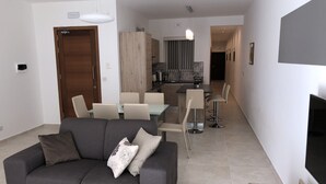 A modern living room with a grey sofa, a wooden dining table, and a kitchen in the background. - Azure Cottage: Beautiful, modern 3 bedroom apartment with communal swimming pool (Marsalforn)