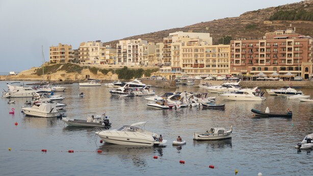 A marina with various boats docked, a building with multiple balconies in the background, and a hilly landscape. - Azure Cottage: Beautiful, modern 3 bedroom apartment with communal swimming pool (Marsalforn)
