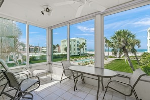 A sunlit balcony with a table and chairs, overlooking a pool and a beachfront view.
