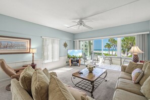 A spacious living room with a large window offering a view of the beach, a ceiling fan, and a flat-screen TV.