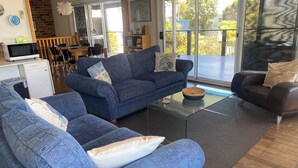 Un salon moderne avec un canapé bleu, une table basse en verre et un fauteuil en cuir foncé. On aperçoit à l’arrière-plan un coin cuisine avec un four à micro-ondes et des placards.