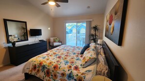 Un dormitorio con cama, tocador, televisor y un balcón con vistas a casas.