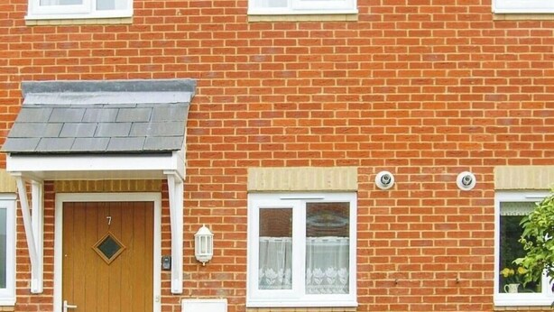 Una casa de ladrillo de dos pisos con puerta principal, un pequeño porche y una ventana con cortina.