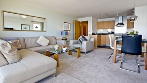 A modern living room with a beige sofa, a glass coffee table, and a dining area with a blue chair and a white table. - Harbour View - A6 Masts (Torquay)