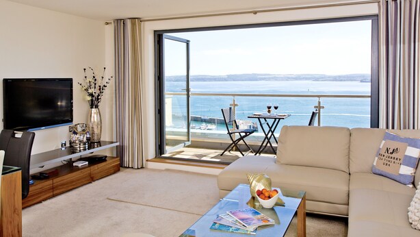 A modern living room with a flat-screen TV, a coffee table with magazines, a sofa with a decorative pillow, and a balcony with a view of the sea. - Harbour View - A6 Masts (Torquay)