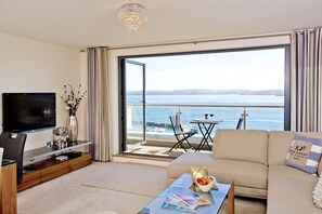 Uma sala de estar moderna com televisão de tela plana, mesa de centro com revistas, sofá com almofada decorativa e varanda com vista para o mar.