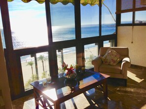 Una habitación con un ventanal grande que da a una playa, una mesa de madera con un florero y velas, y un sillón color beige con un cojín estampado.