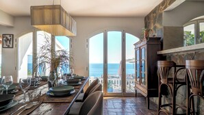 Un coin repas avec vue sur la mer, une table en bois dressée avec des assiettes et des verres, et un luminaire suspendu.