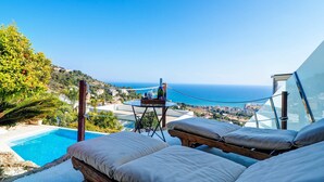 Un espace aménagé au bord de la piscine, avec des chaises longues, une table sur laquelle est posée une bouteille, et une vue sur la mer et la ville.