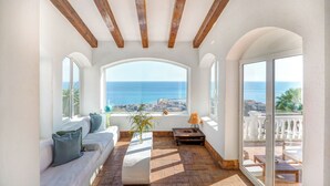 Un intérieur lumineux, avec un canapé blanc, une petite table et une vue sur la mer grâce à de grandes fenêtres.