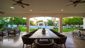 Une terrasse extérieure avec une longue table et des chaises, une piscine en arrière-plan et des ventilateurs de plafond.