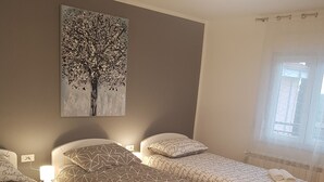 Ett hotellrum med tre sängar, en stor tavla med ett trädmotiv på väggen och ett fönster med gardiner.