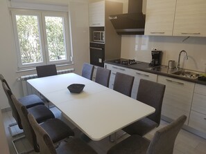 A modern kitchen with a white dining table, grey chairs, a built-in oven, and a window with a view of trees.