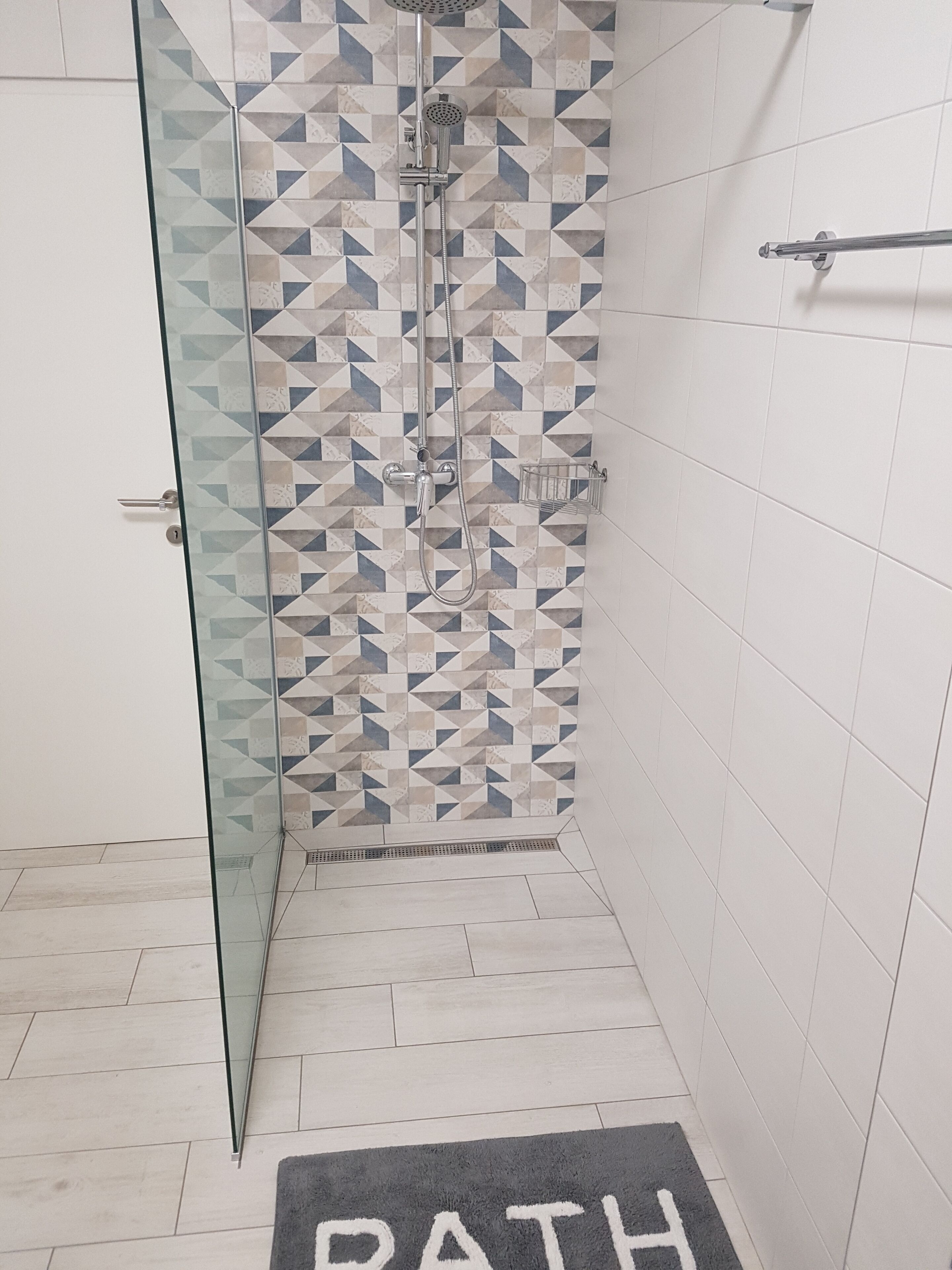 A shower area with a geometric tile pattern, a glass door, and a "RATH" rug on the floor.