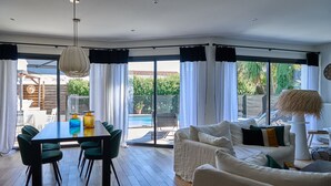 A spacious living area with a dining table, green chairs, a white sofa, and a large window offering a view of a swimming pool and greenery. - Bruges, Near Bordeaux: Charming, Air-Conditioned House with a Pool and Garden (Bruges proche Bordeaux)