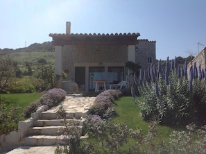 Un bâtiment en pierre avec un toit en tuiles, un escalier menant à l’entrée et un jardin agrémenté de fleurs violettes.