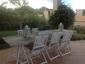 Une table à manger rustique, installée à l’extérieur, avec des chaises blanches, entourée de verdure et avec un bâtiment en pierre en arrière-plan.