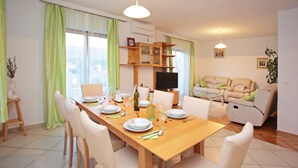 Un coin repas avec une table en bois dressée pour quatre personnes, entourée de chaises blanches. On aperçoit un canapé et un téléviseur au fond.