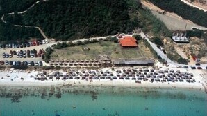 An aerial view of a coastal area with a large parking lot, a building with a red roof, and a beach with numerous cars.