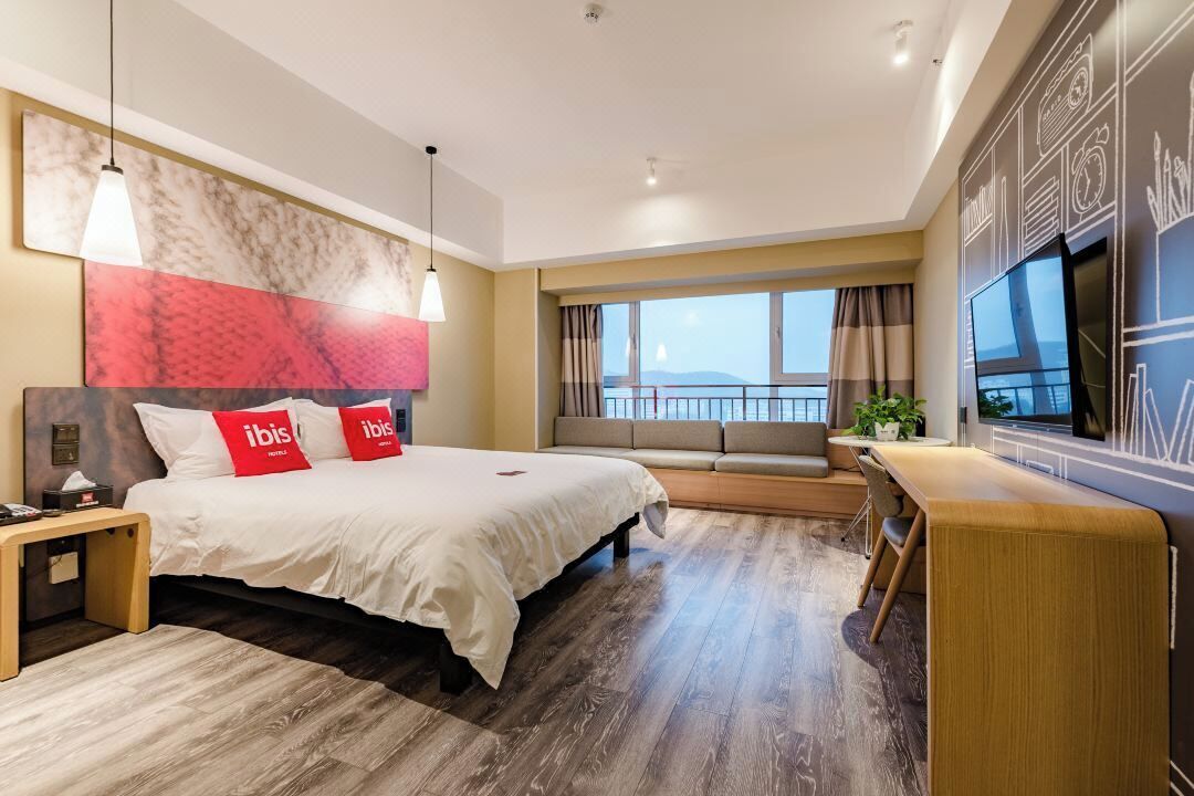 A modern hotel room with a large bed, a desk, a TV, and a view of the sea.