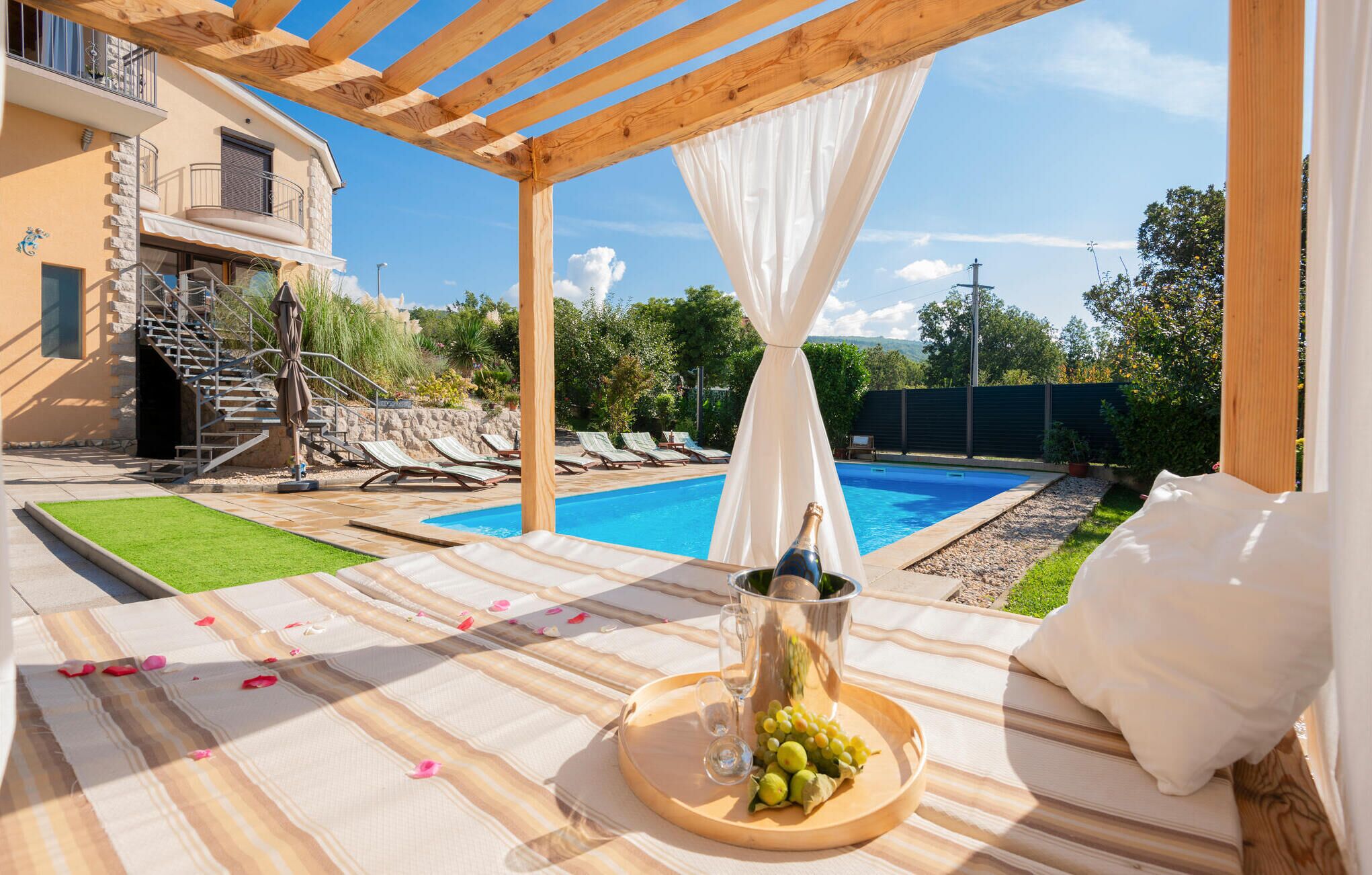 A poolside setting with a pergola, a bottle of champagne, and a fruit platter on a table.