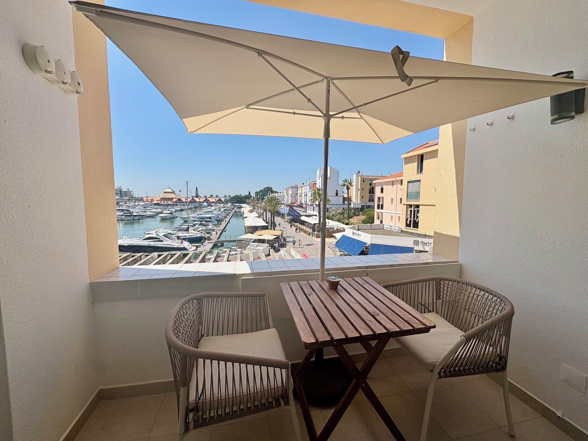 Un balcon avec une table et deux chaises, donnant sur une marina où l’on peut admirer des bateaux et des bâtiments.