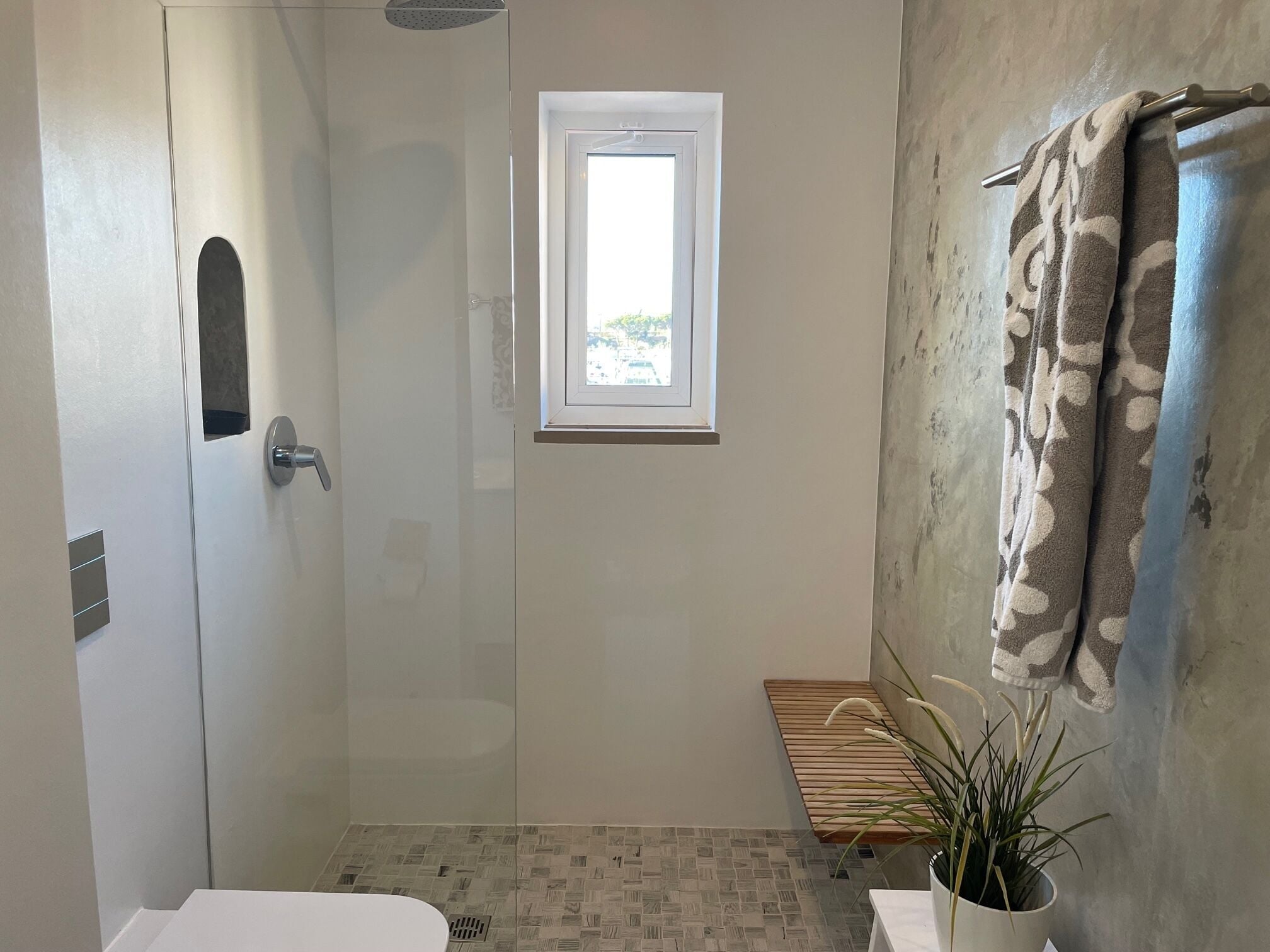 A modern bathroom with a shower, a small window, a bench, and a potted plant.