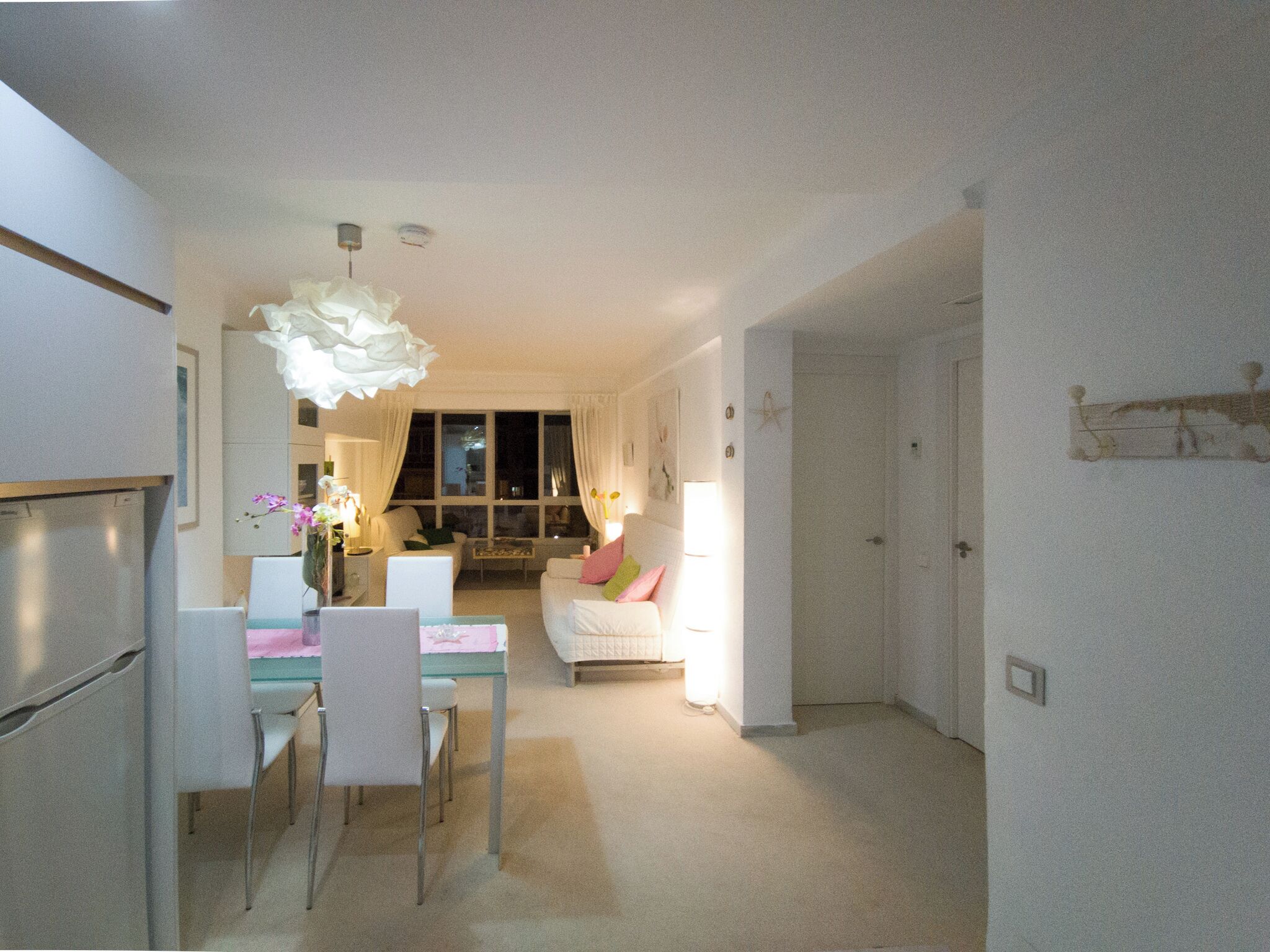 Une cuisine moderne avec des meubles blancs, une table à manger avec des chaises et un lustre. Le salon est doté d’un canapé blanc et d’une table basse.