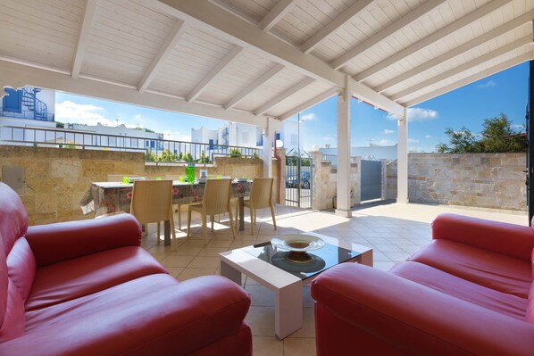 A covered outdoor seating area with red sofas and a table, a dining table with chairs, and a view of the cityscape.