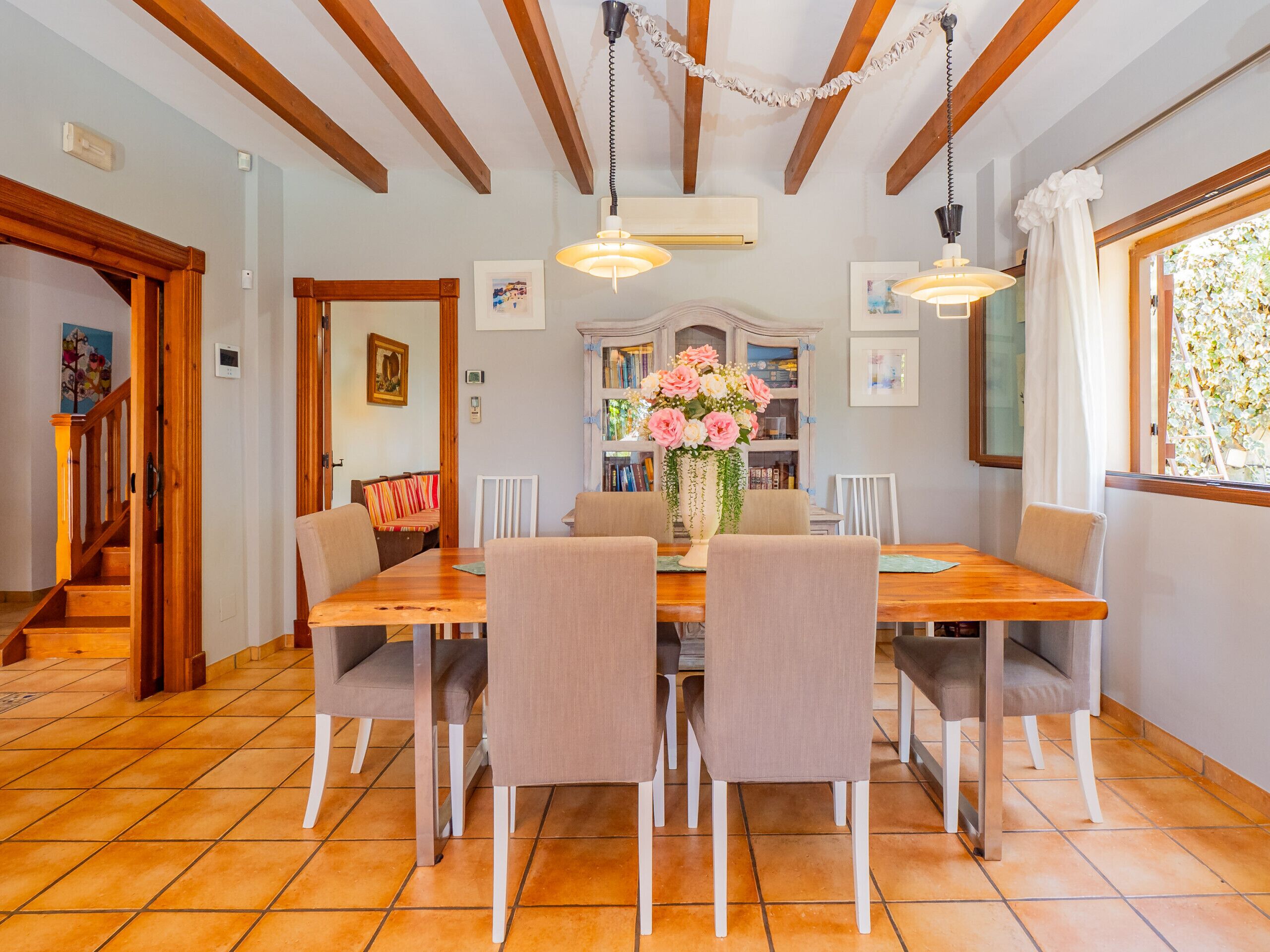 A dining area with a wooden table and chairs, a vase of flowers, and a view of the garden through a window.