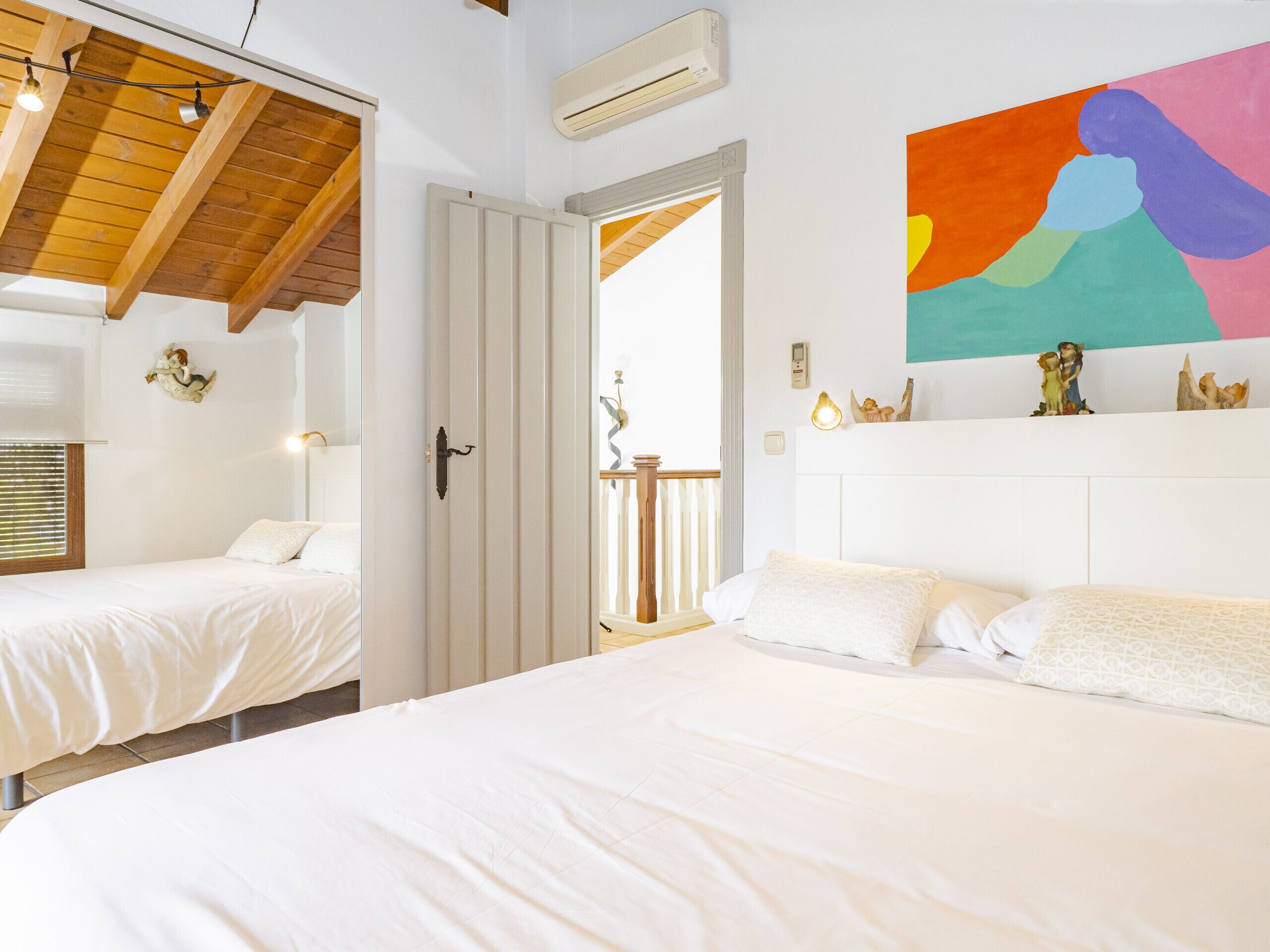 Ett hotellrum med två sängar, ett träpanelat tak, en luftkonditioneringsanläggning och en färgstark, abstrakt målning på väggen.