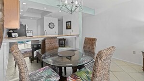 A modern dining area with a glass table surrounded by wicker chairs, a geometric chandelier, and a kitchen area with a Wi-Fi sign. - Gulfside Comfort – Beach Castle #4 by RVA (Longboat Key)