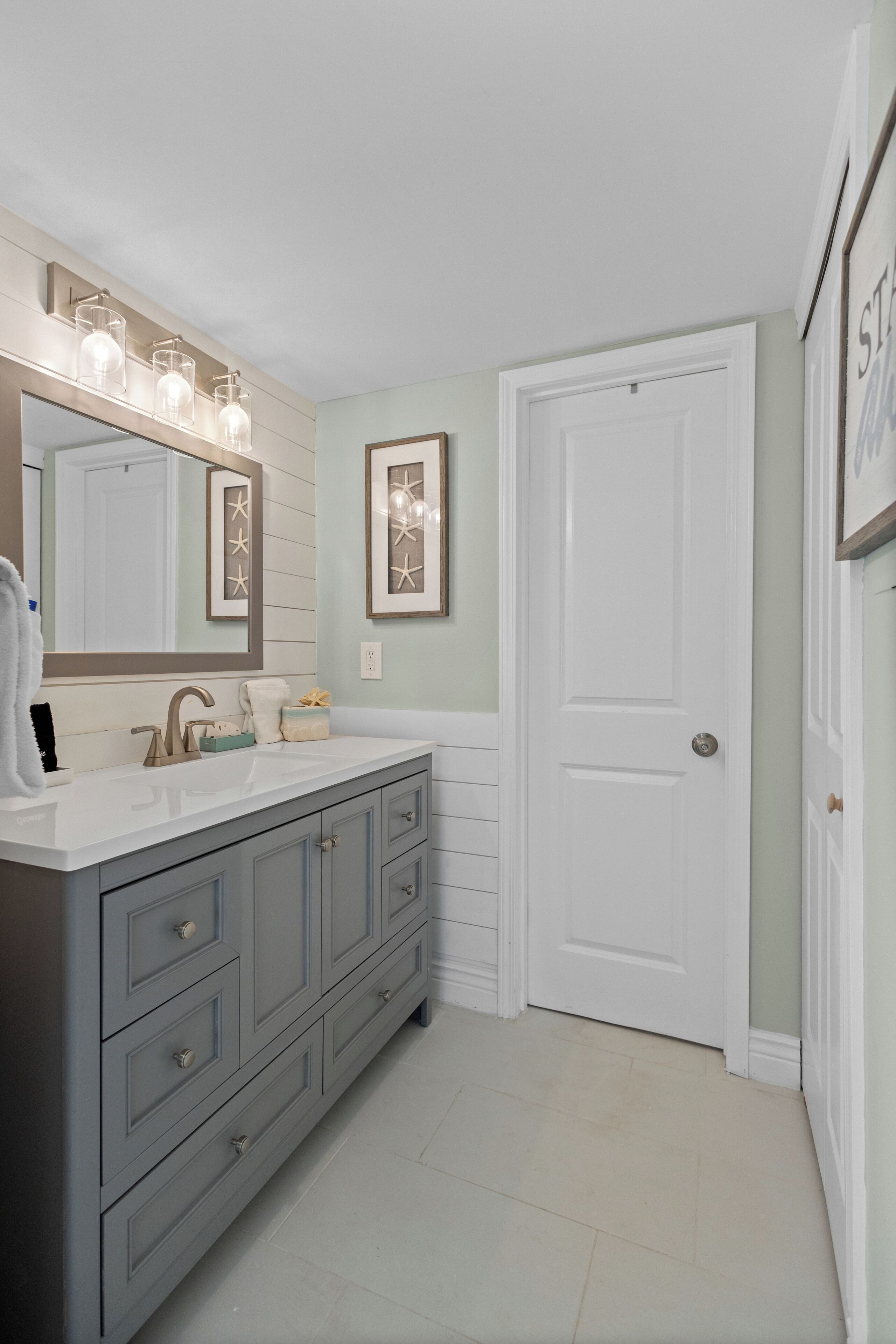 A bathroom with a double vanity, a large mirror with lights, and a white door.