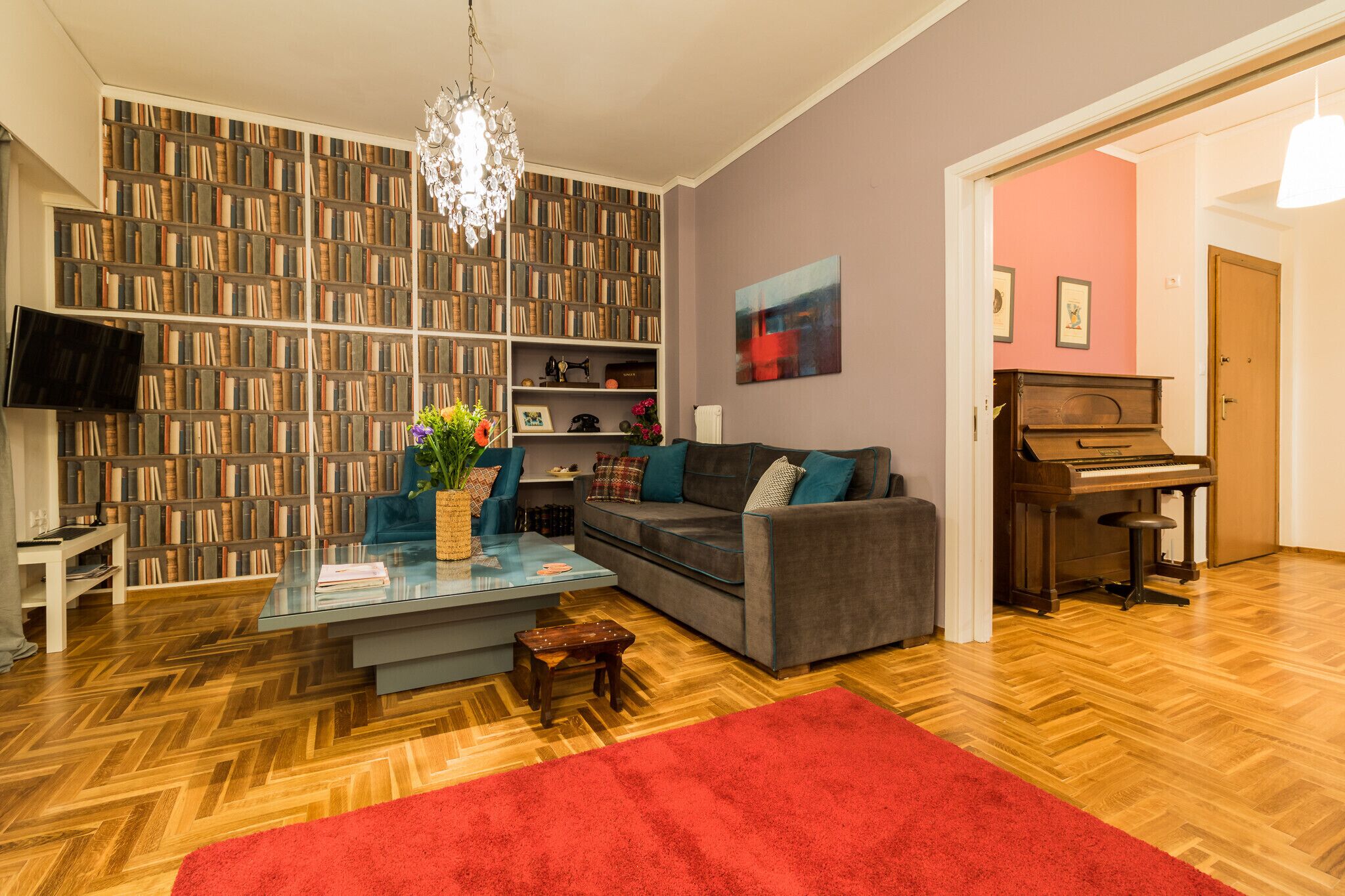 Een woonkamer met een boekenkast, een bank, een salontafel, een rood kleed en een piano.