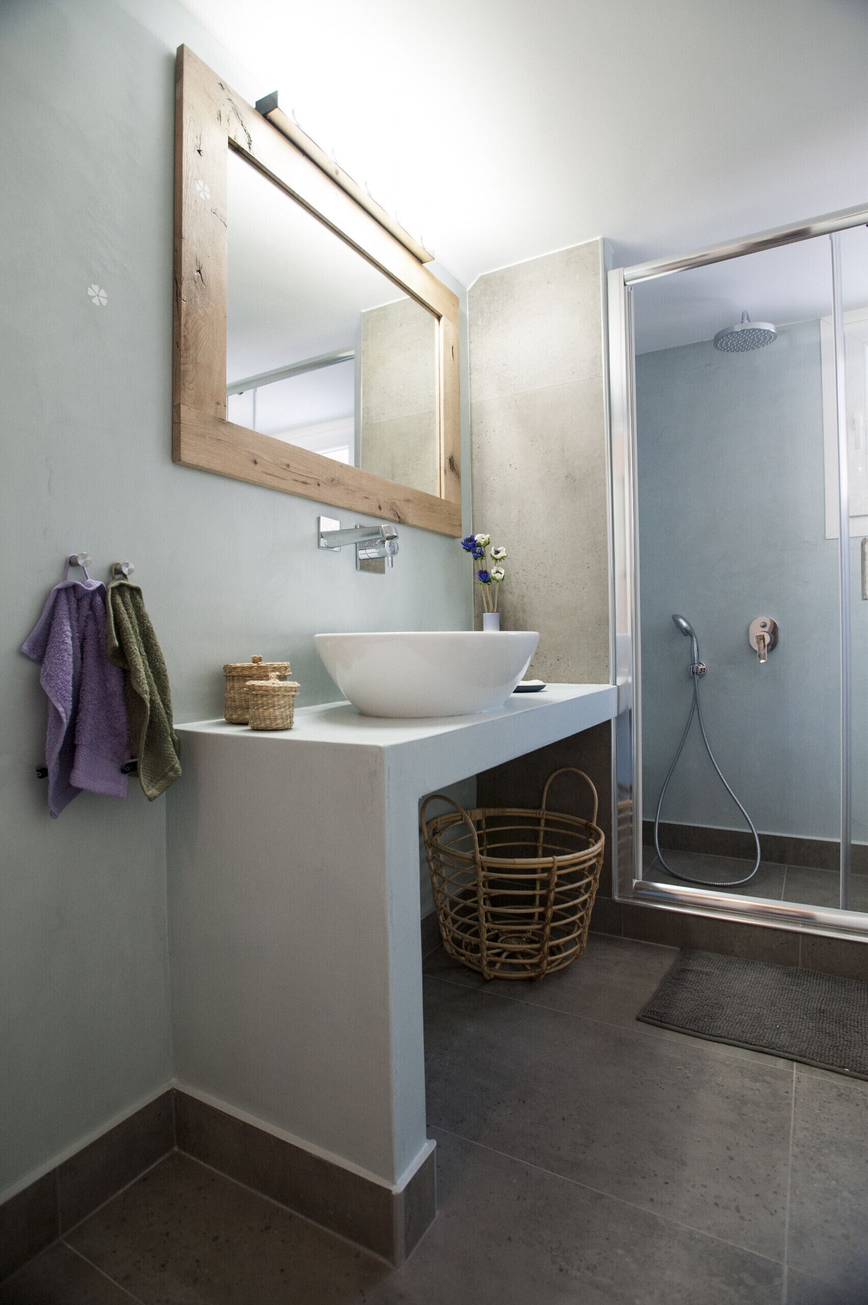 Une salle de bain avec un lavabo blanc, un miroir à cadre en bois, une douche et un panier en osier.