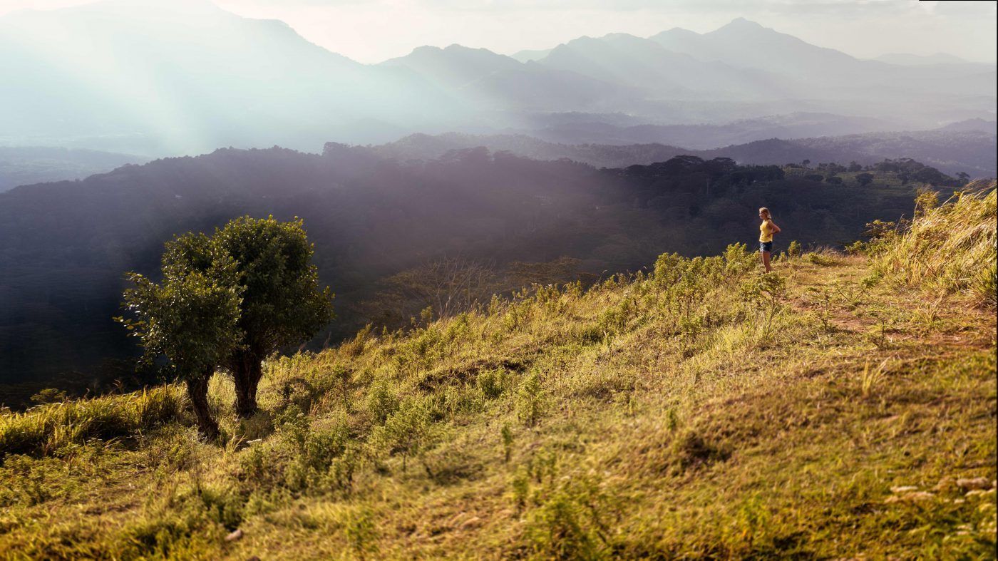 A hilly landscape with a person standing on a grassy slope, overlooking a valley with distant mountains.