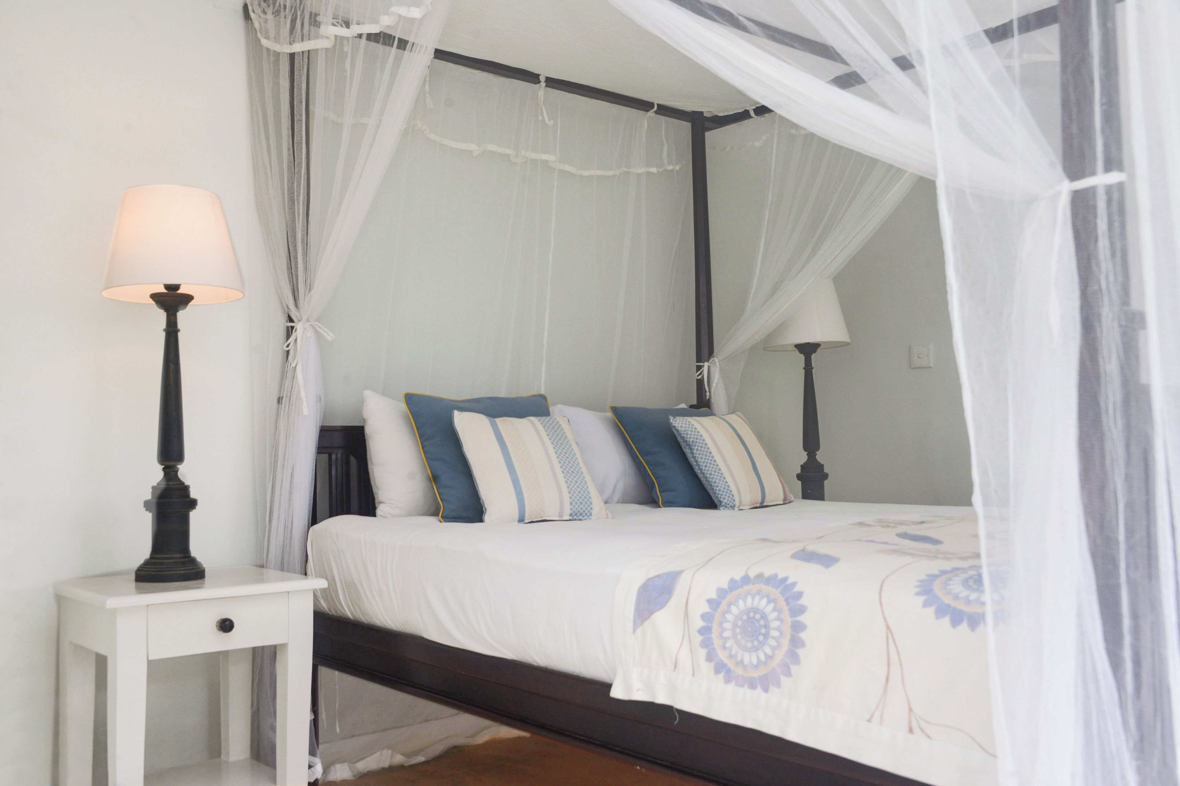Quarto com cama de dossel, roupa de cama branca e mesa de cabeceira com abajur.