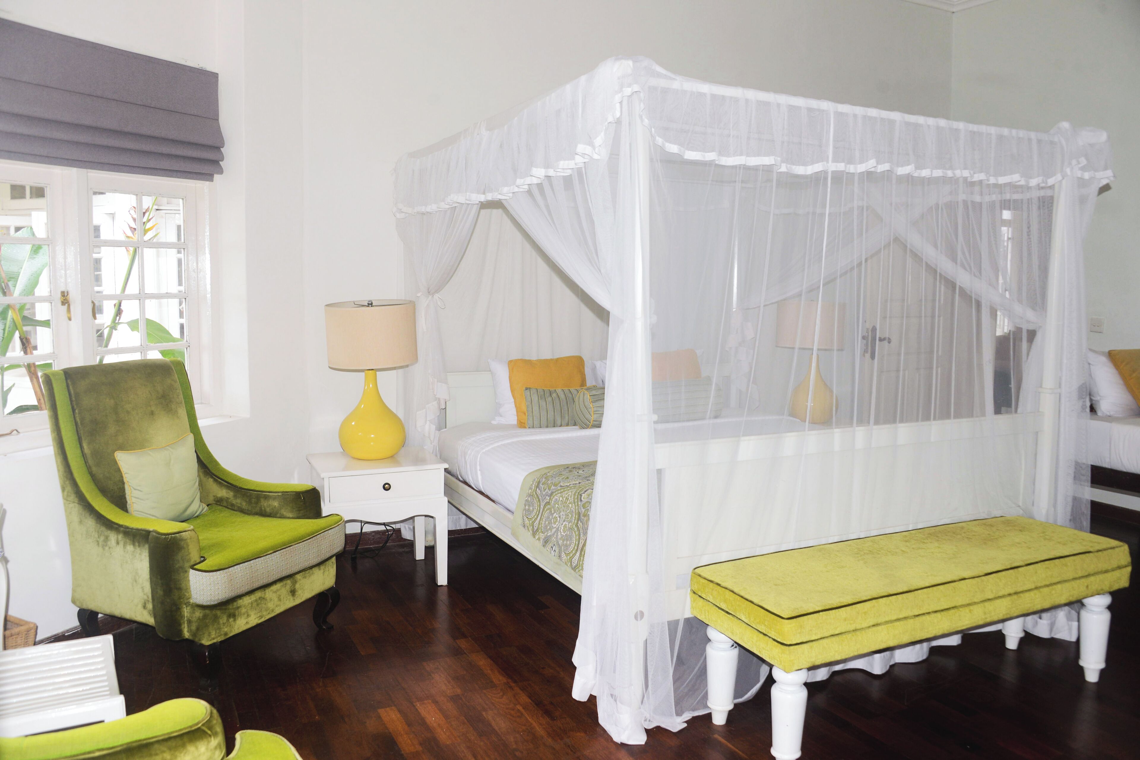 A bedroom with a four-poster canopy bed, a green armchair, a bedside table with a lamp, and a bench.