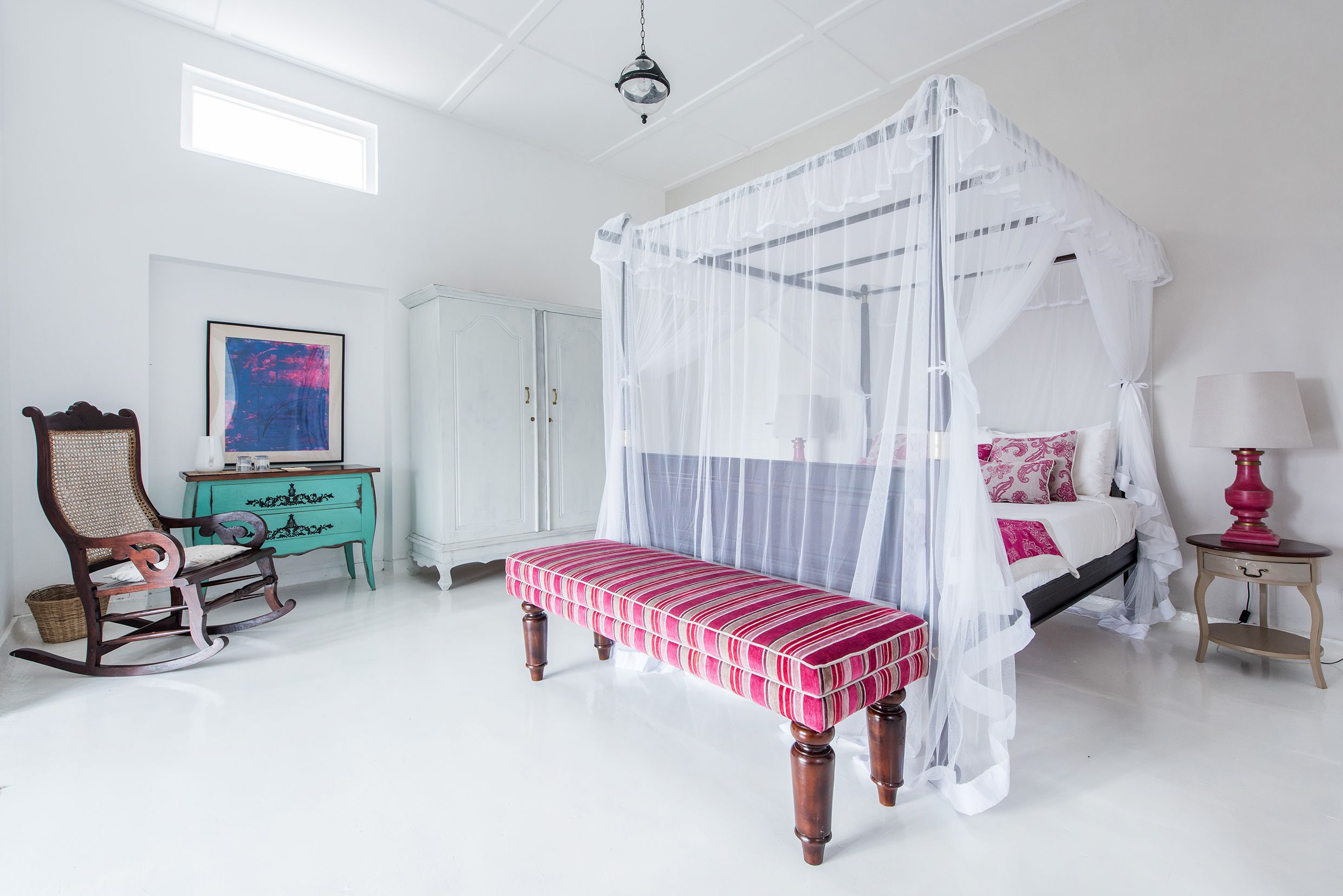 A bedroom with a four-poster bed, a rocking chair, a bench, a turquoise dresser, and a framed picture on the wall.