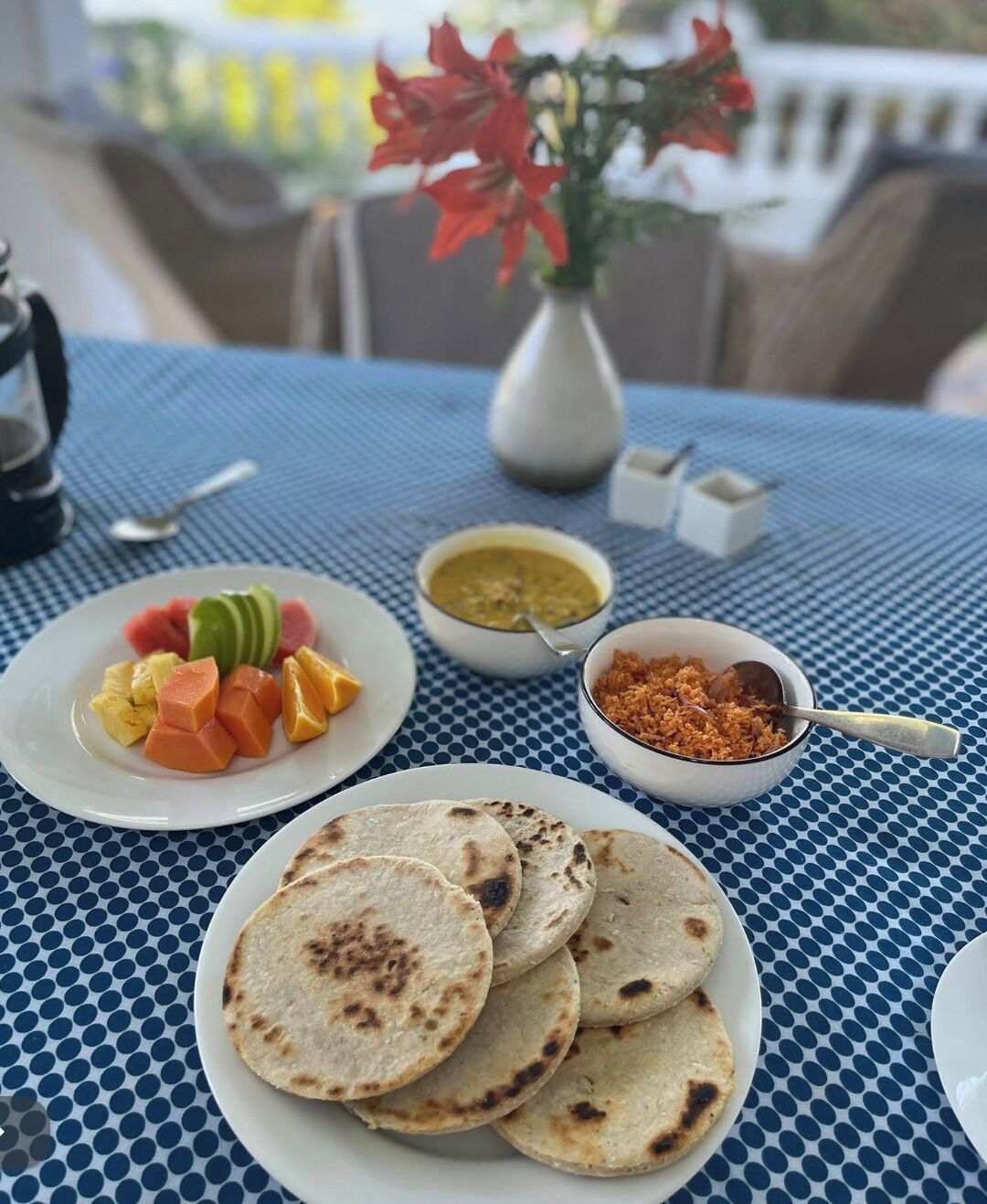 A table with a blue checkered tablecloth, a white plate with sliced fruits, a bowl of curry, a bowl of brown sugar, and a white vase with red flowers.