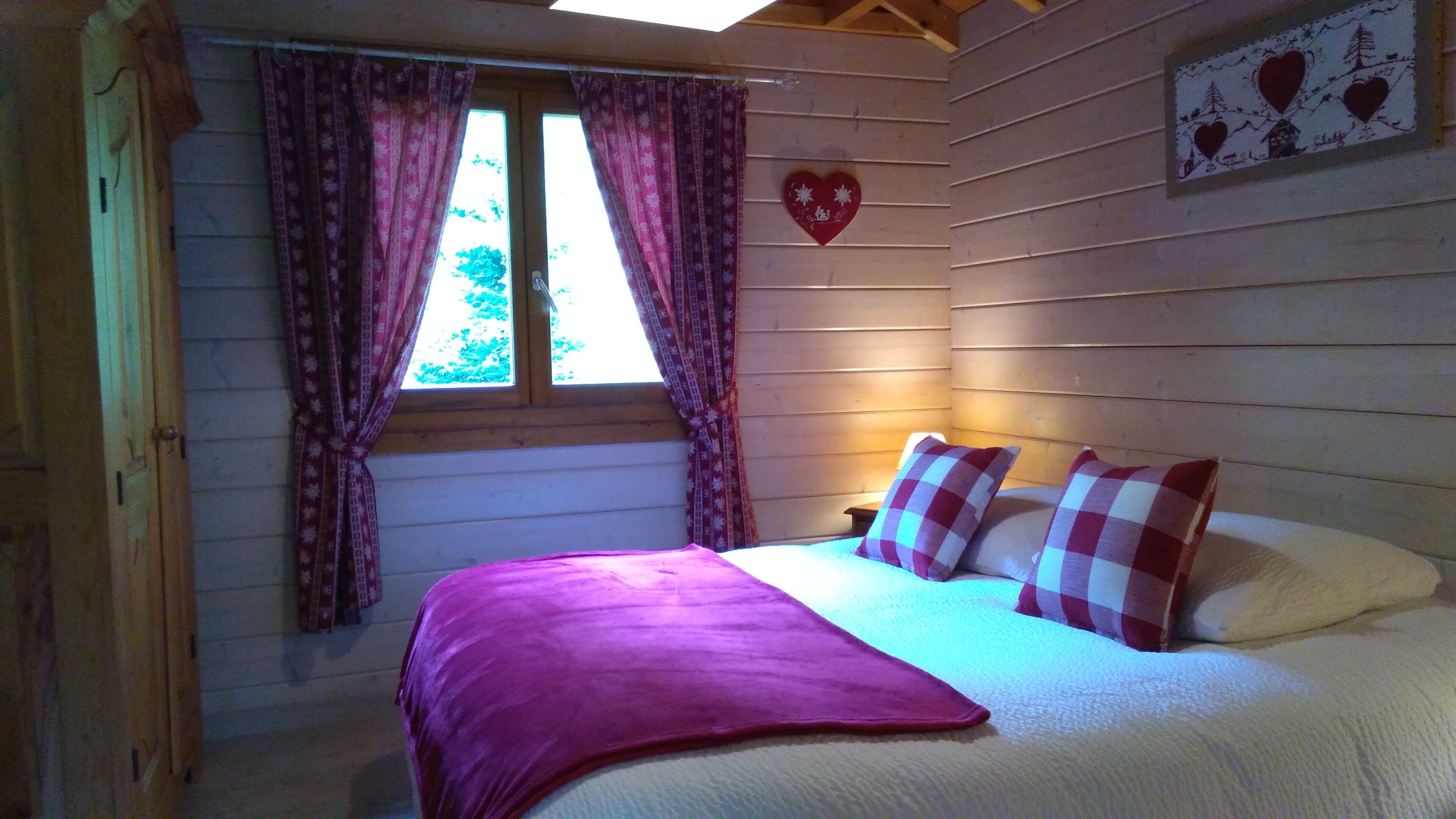 A cozy bedroom with a bed, a window with curtains, and a heart-shaped decoration on the wall.