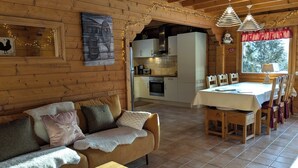A cozy cabin interior with a wooden dining table, chairs, a sofa with cushions, a kitchen area, and a painting on the wall.